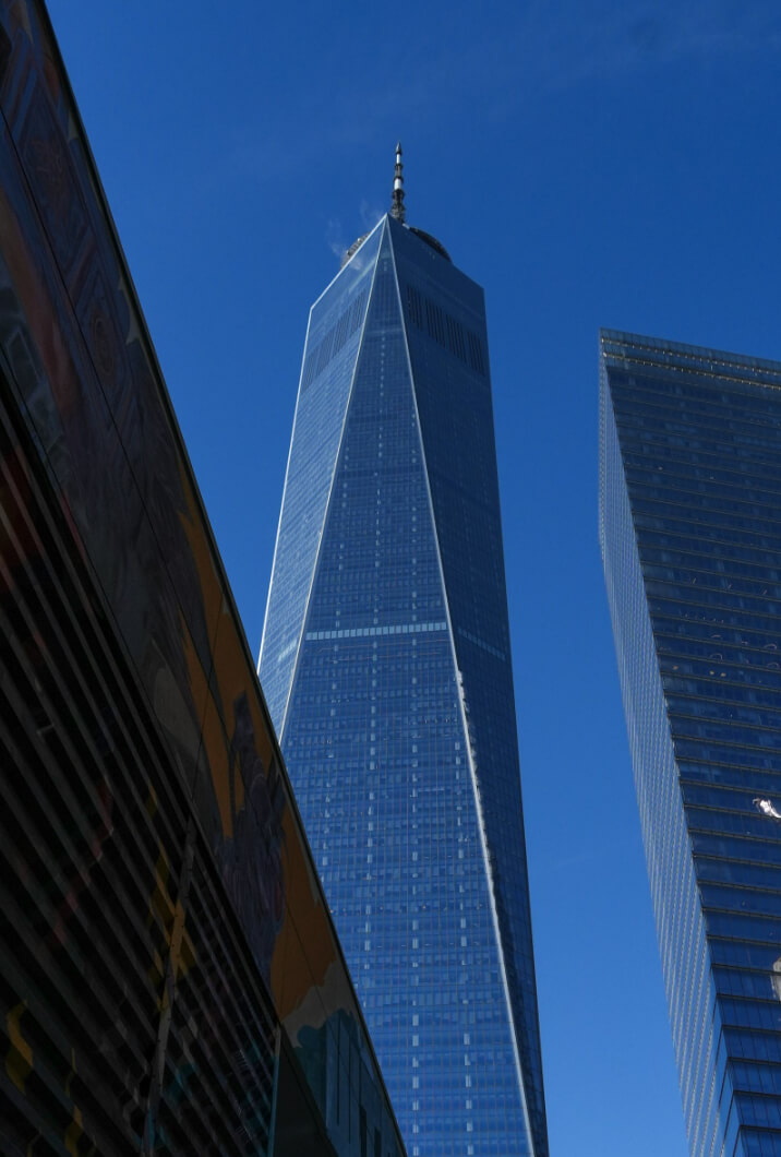 View of a tall, narrow skyscraper with a pointed top against a blue sky, flanked by other modern buildings.