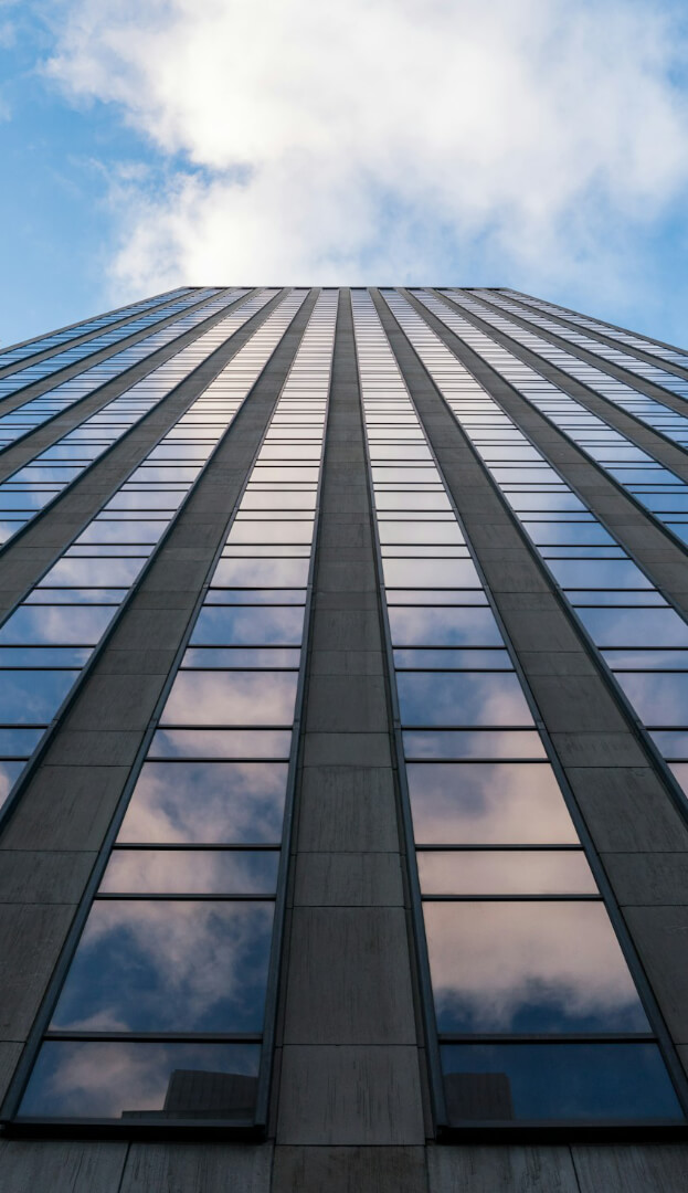 A tall skyscraper with a glass facade reflects clouds in the sky, shot from a low angle looking upwards.