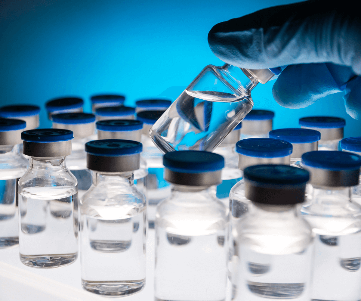 A gloved hand holding a vial above several rows of similar vials filled with clear liquid, all with blue caps, against a blue background.