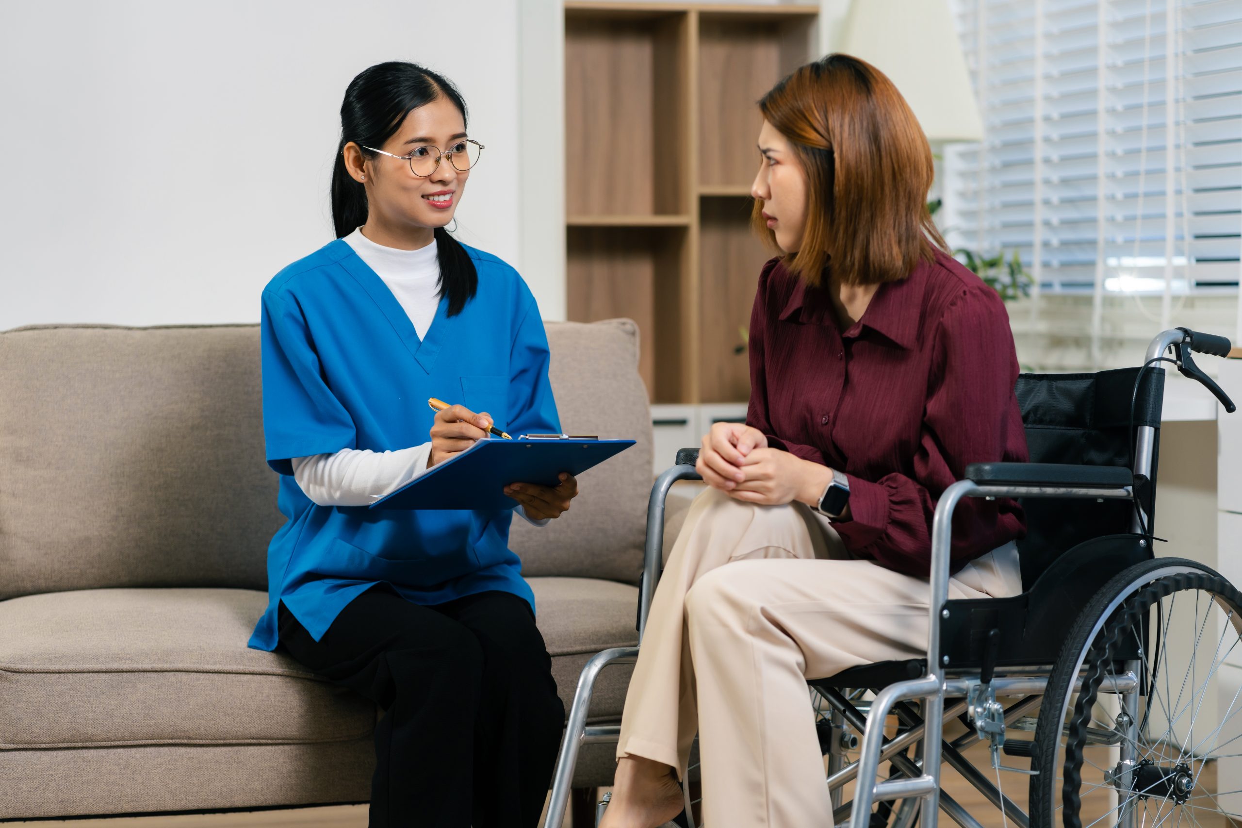 A healthcare worker with a clipboard talks to a seated woman in a wheelchair in a living room setting.