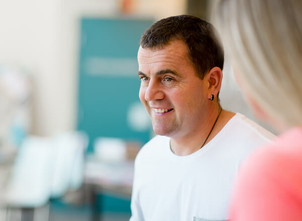 A man with short dark hair and an earring smiles while talking to a person with long blonde hair; indoor setting with blurred background.