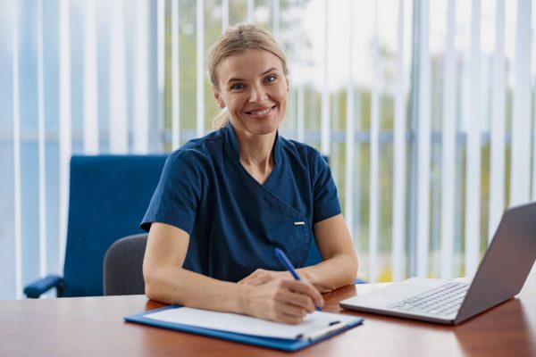 A woman in medical scrubs sits at a desk, smiling, while writing on a clipboard next to an open laptop.