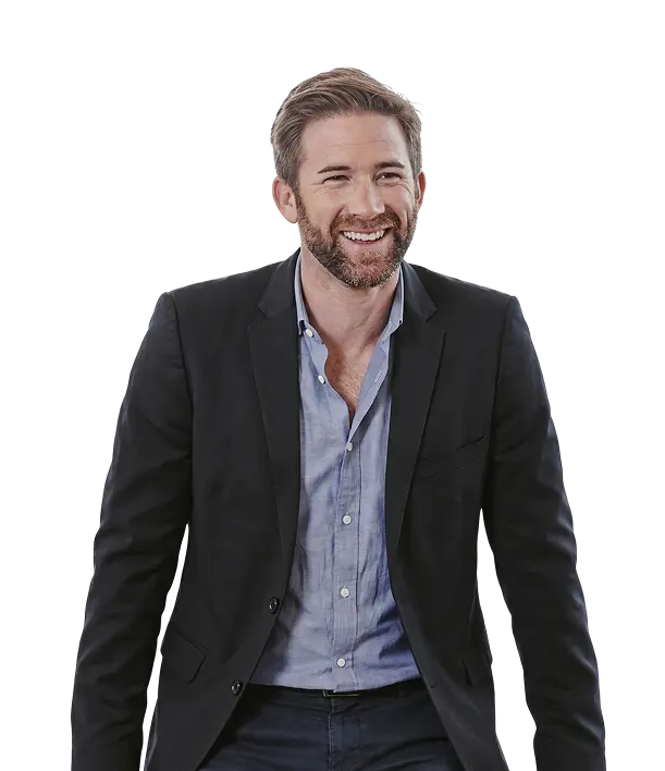 A man with light brown hair and a beard is wearing a dark blazer over a light blue shirt, smiling and looking slightly to the side.