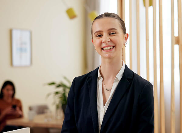 A woman in a dark blazer and white blouse smiles while standing indoors; a blurred figure is seated in the background using a phone.