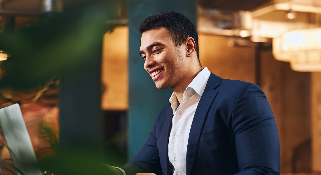 A man in a business suit sits at a table, smiling while looking at a laptop in a modern indoor setting.