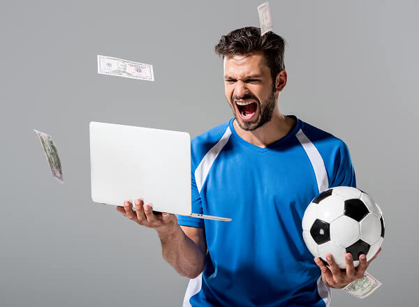 Man in blue sports shirt holding a soccer ball and laptop, appearing excited, with paper money falling around him against a gray background.