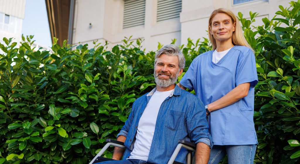 A man sits in a wheelchair outdoors while a woman in blue scrubs stands beside him, both looking at the camera in front of green bushes and a building.