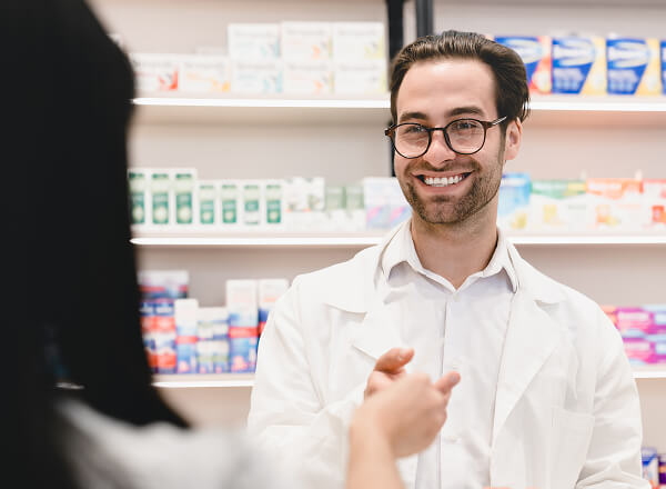 A pharmacist in a white coat smiles while handing a prescription to a customer at a pharmacy counter with shelves of medicine in the background.