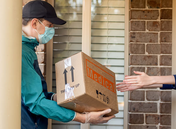 A delivery person wearing a mask hands a box labeled "Medical" to someone at a doorstep.