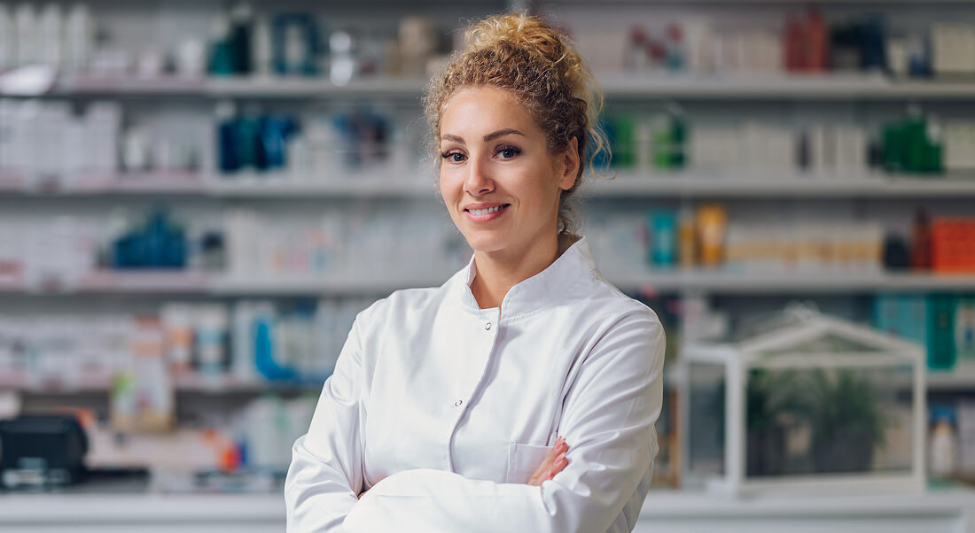 A woman in a white lab coat stands with arms crossed, smiling, in front of shelves stocked with pharmaceutical products in a pharmacy.