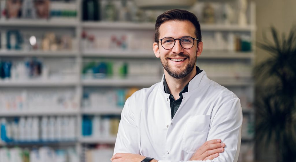A man in a white lab coat and glasses stands smiling with arms crossed in a pharmacy, shelves of medical products visible in the background.