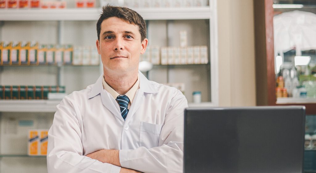 A pharmacist in a white coat stands with arms crossed behind a counter in a pharmacy, with shelves of medication visible in the background.