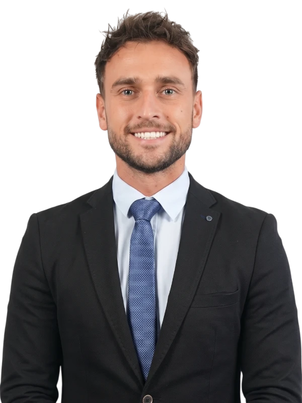 Michael Barrett in a black suit, light blue shirt, and blue tie is standing and smiling at the camera against a plain background.