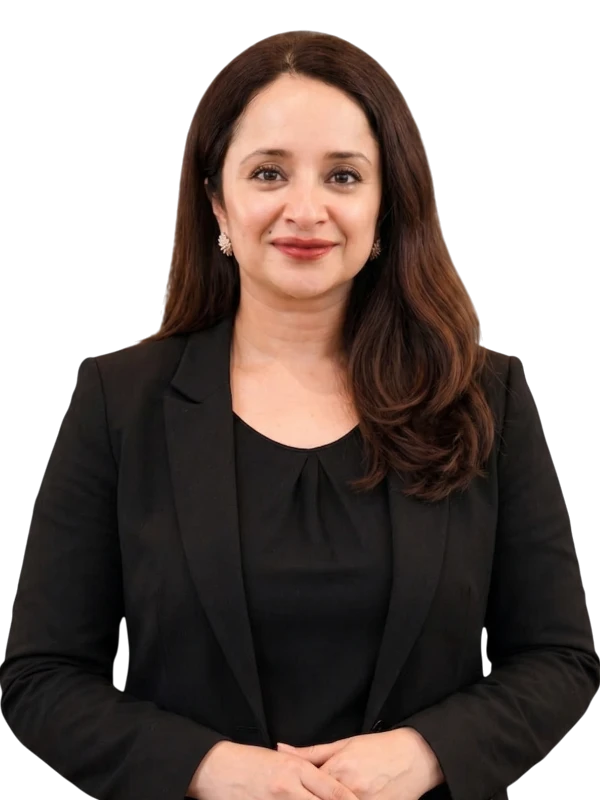 Reema Alwadhi wearing a black blazer and black top stands facing the camera, smiling with her hands clasped in front of her.