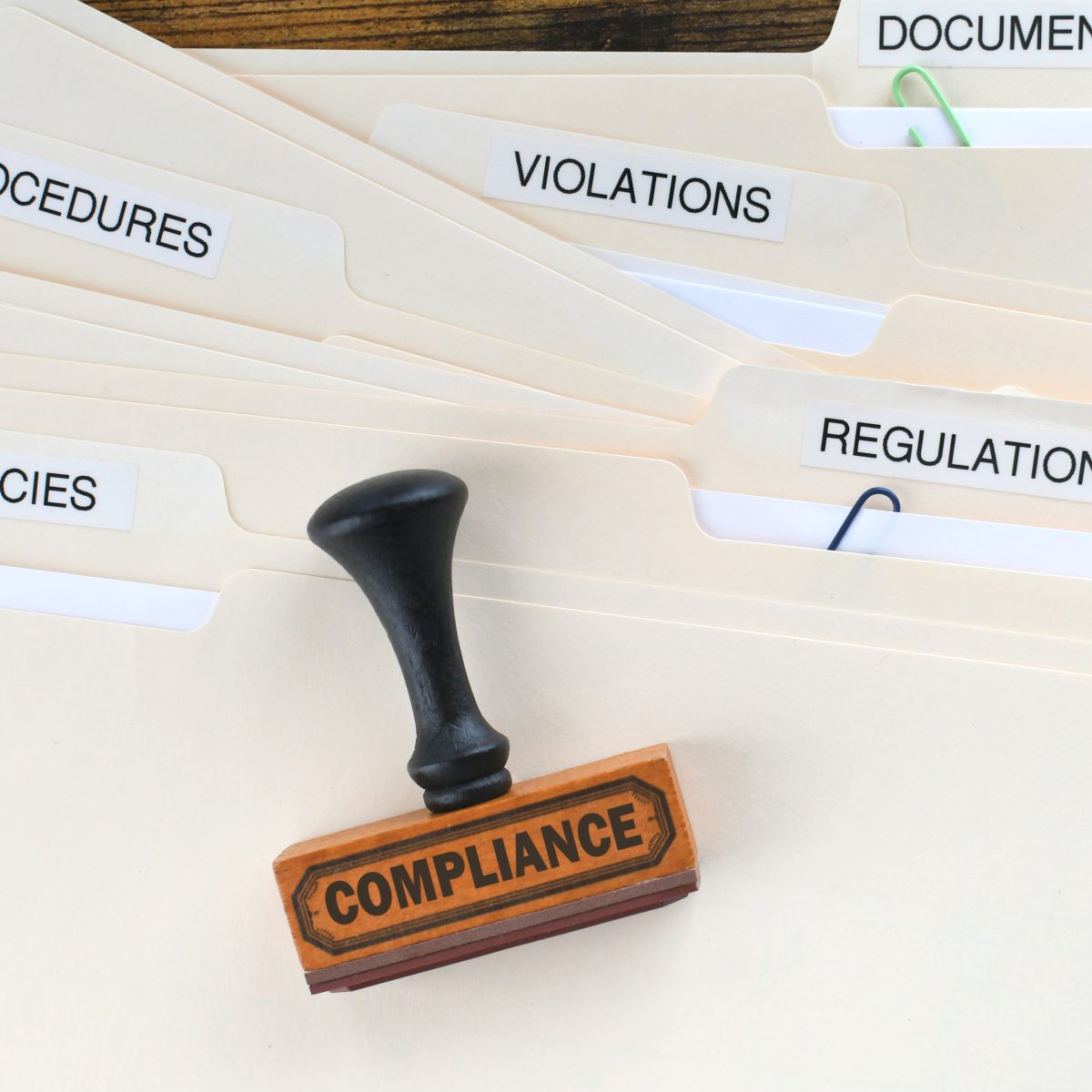 Folders labeled documentation, procedures, violations, regulations, and policies with a rubber "COMPLIANCE" stamp placed on top of them.