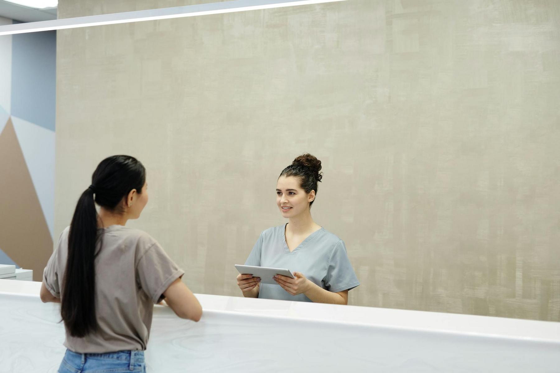 A receptionist in scrubs holds a tablet and talks to a woman standing at the front desk in a modern office or clinic.