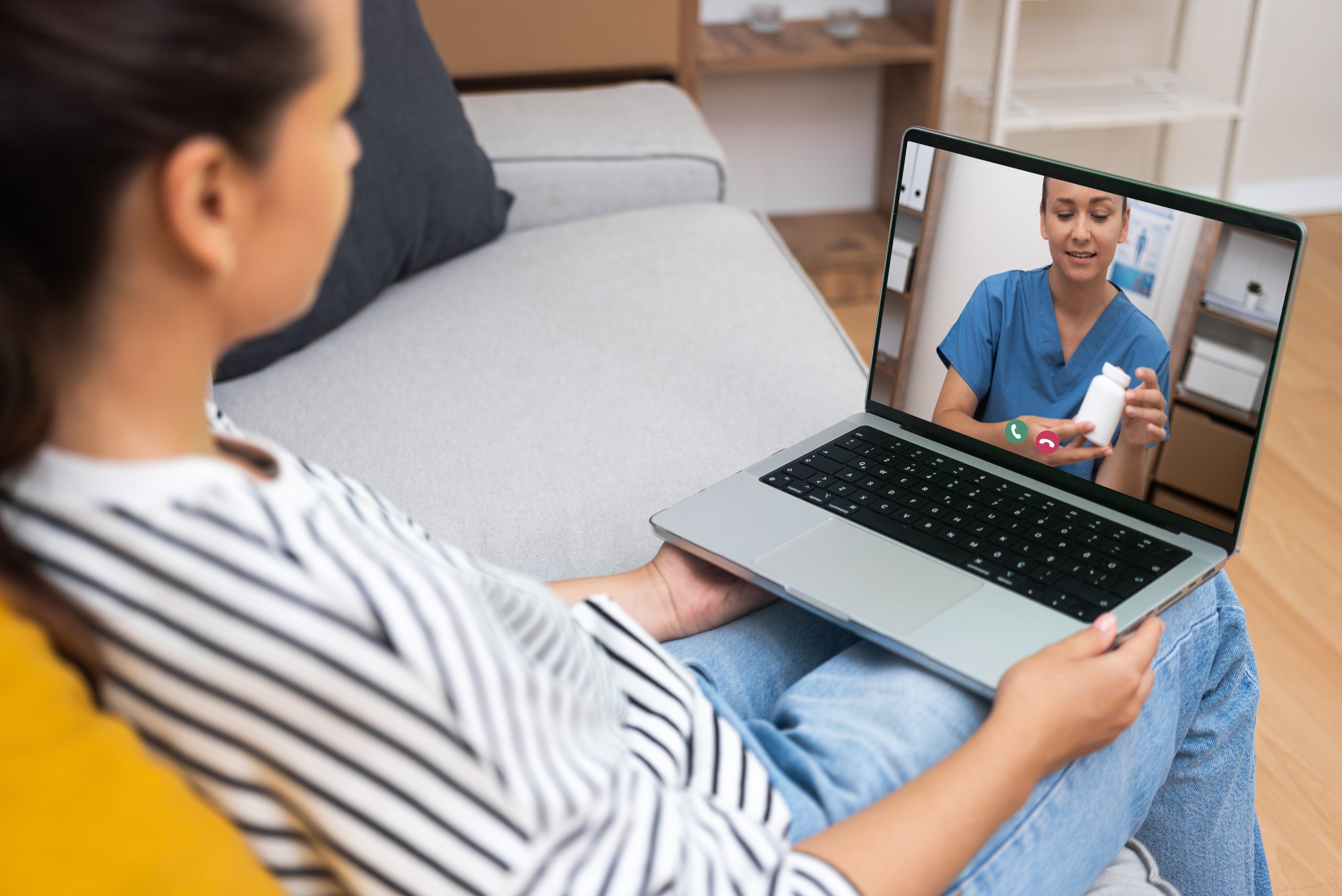 A woman sits on a couch holding a laptop, having a video call with a healthcare professional who is demonstrating medication.