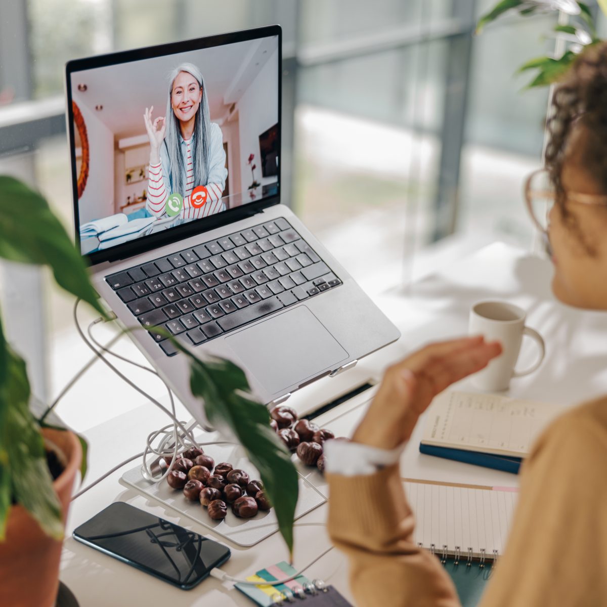 A person participates in a video call on a laptop at a desk with a notebook, pen, phone, and potted plant.