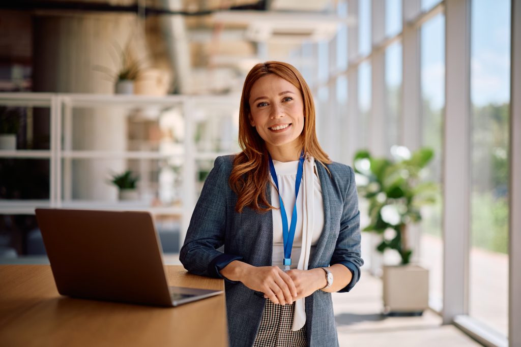 A woman with long reddish hair stands in a modern office space next to an open laptop, smiling at the camera and wearing a blazer with a lanyard.
