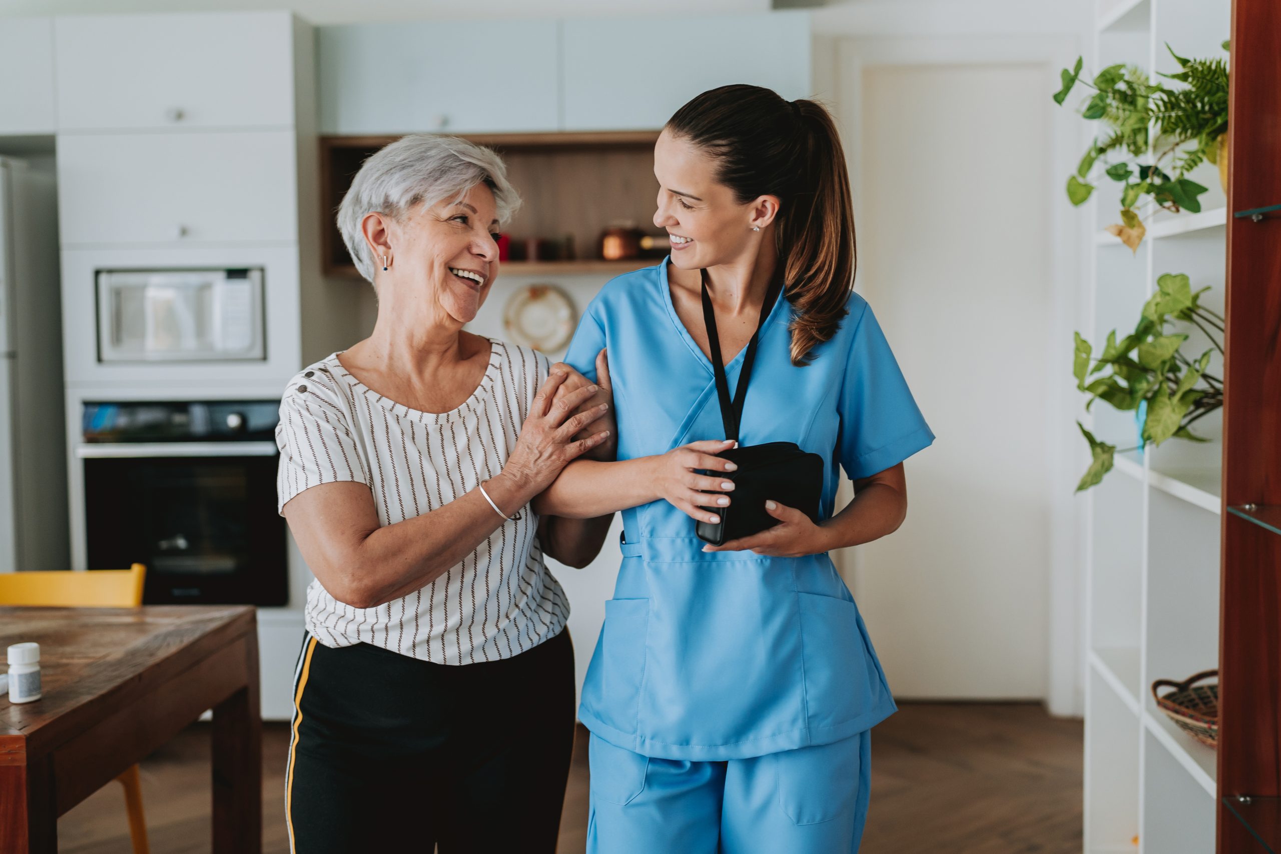 An elderly woman smiles while holding the arm of a healthcare worker in blue scrubs, showcasing the warmth and support provided by aged care services inside a kitchen.