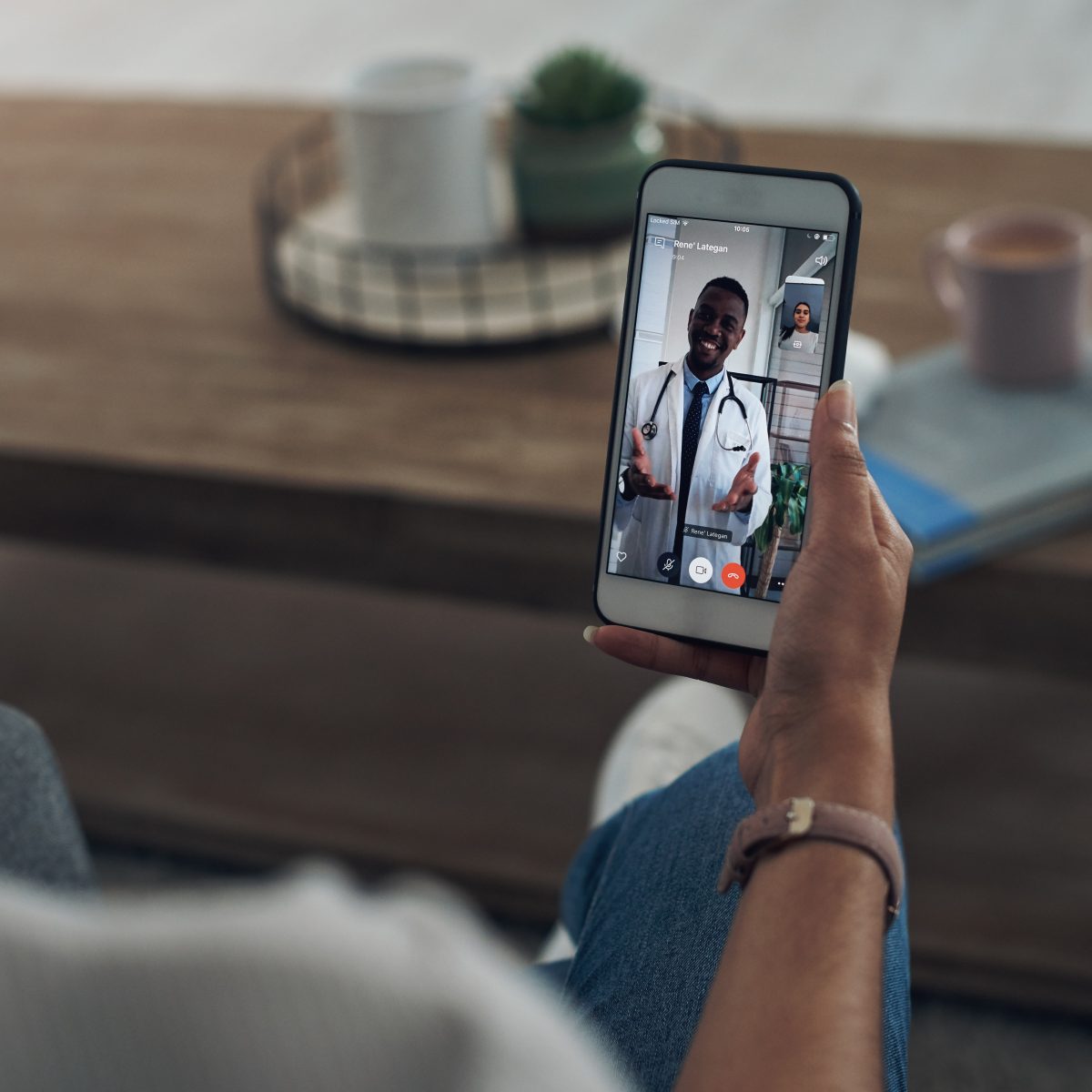 A person holds a smartphone showing a video call with a doctor in a white coat and stethoscope. A coffee table with a mug and plant is in the background.