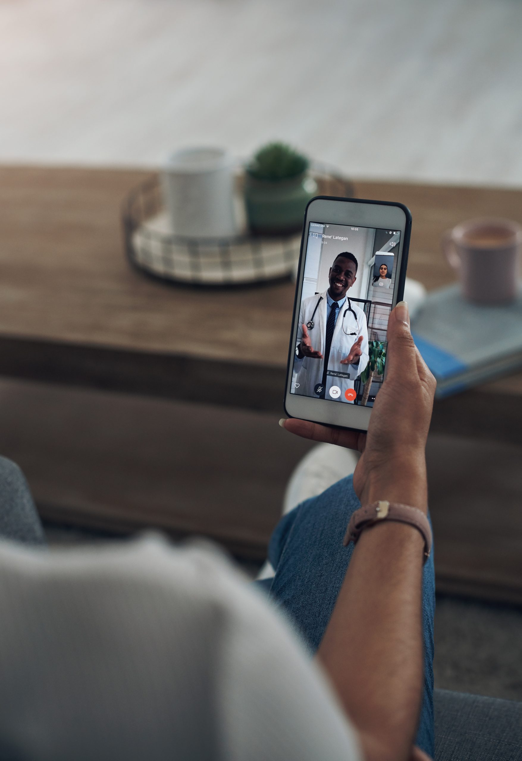 A person holds a smartphone showing a video call with a doctor in a white coat and stethoscope. A coffee table with a mug and plant is in the background.