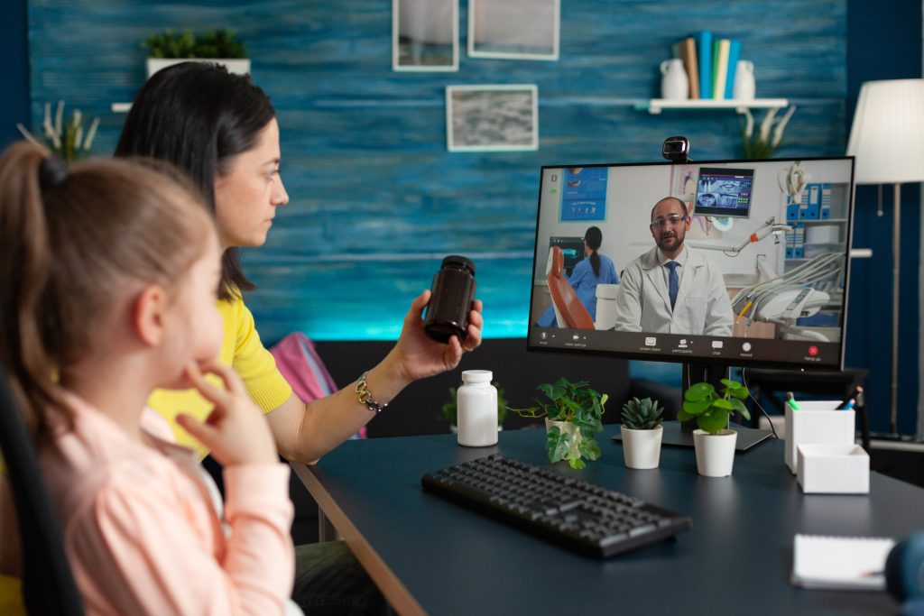 A woman and girl have a telehealth video call with a doctor, showing a medicine bottle; computer monitor and medical items are on the desk.