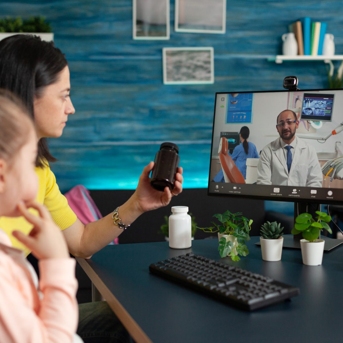 A woman and girl have a telehealth video call with a doctor, showing a medicine bottle; computer monitor and medical items are on the desk.