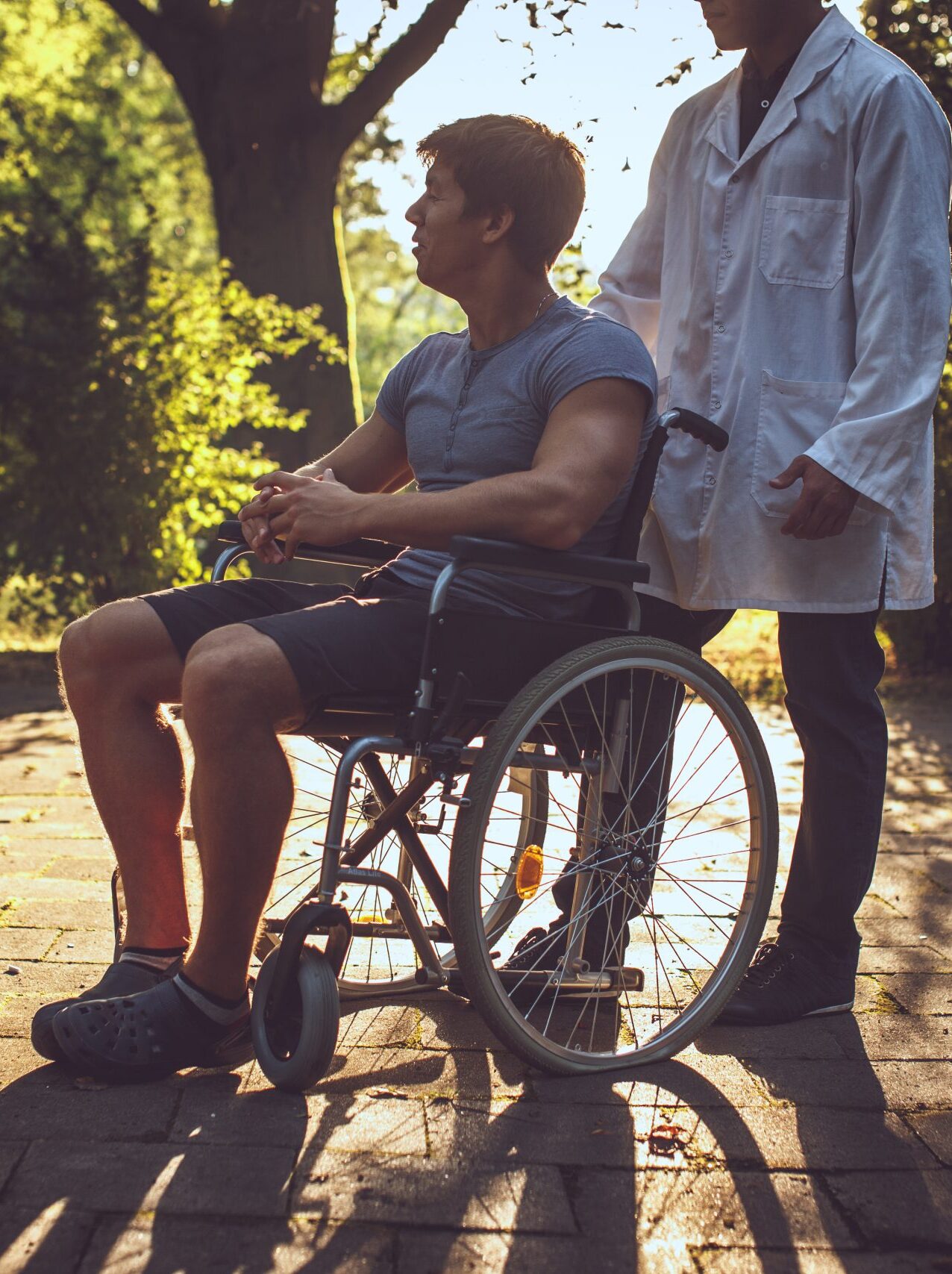 A man in a wheelchair is outdoors on a pathway, accompanied by a person in a white coat. As sunlight filters through the trees, they discuss topics like NDIS certification vs verification.