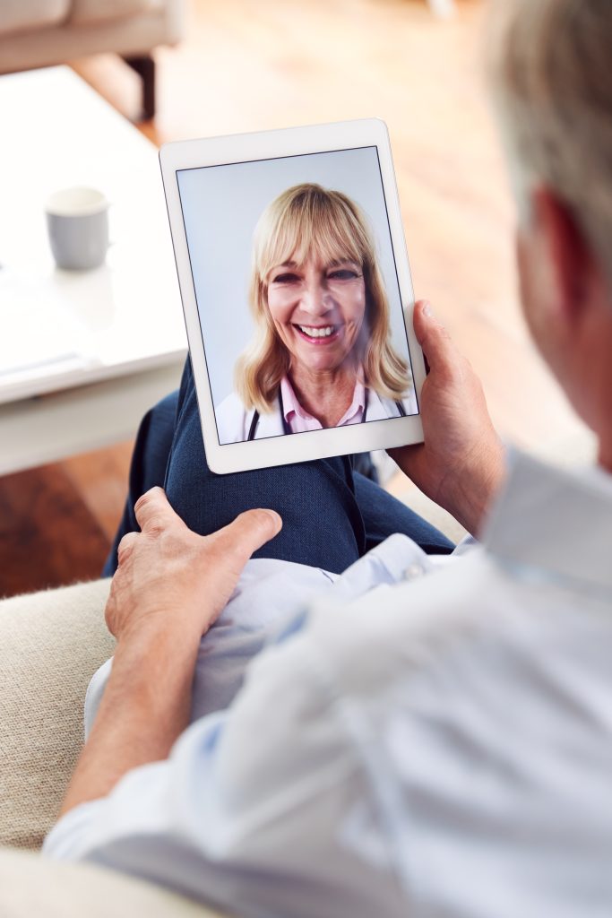 Person holding a tablet showing a woman smiling on a video call, likely a telehealth or virtual consultation in a home setting.