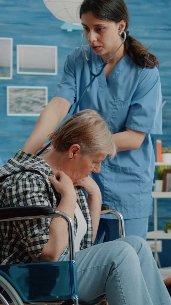 A nurse in blue scrubs uses a stethoscope to check the back of an elderly woman sitting in a wheelchair.
