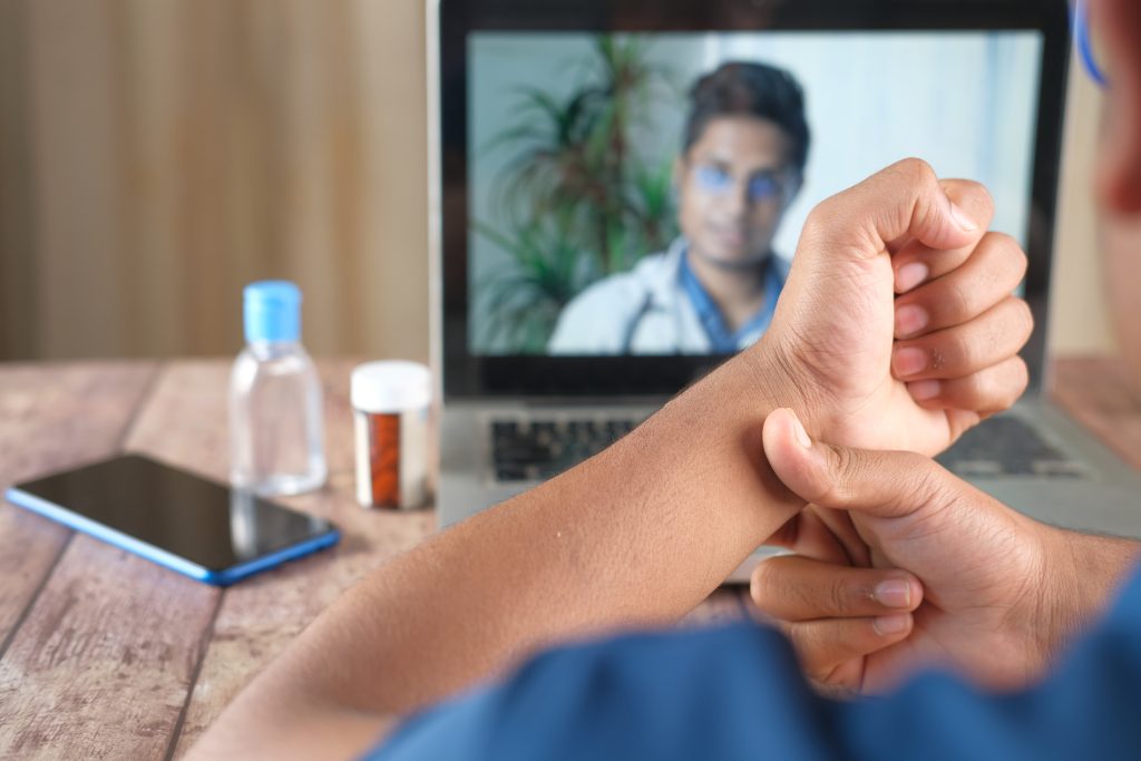 A person checking their pulse while having an online video consultation with a doctor on a laptop; medications and a smartphone are on the table.