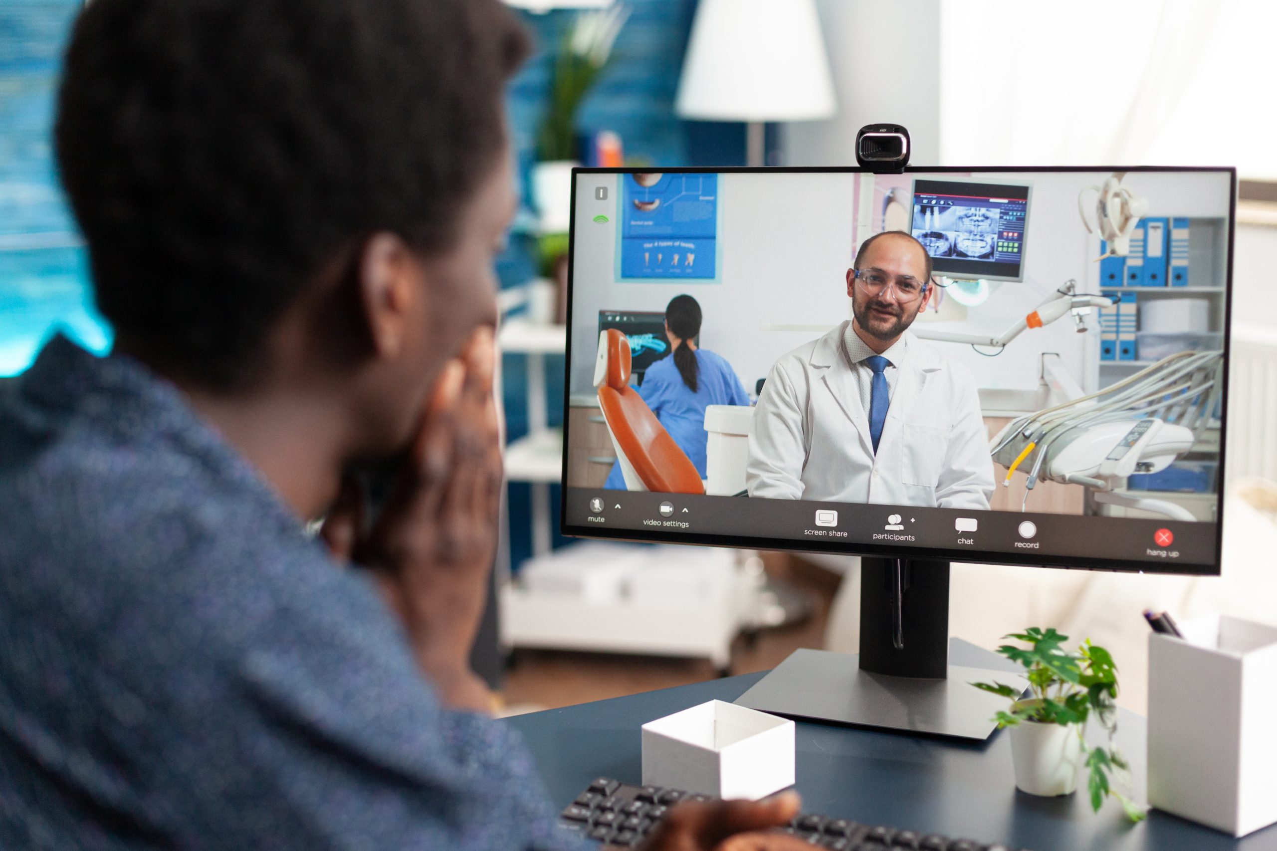 A person has a video call with a doctor, who is sitting in a dental office, using a desktop computer in a home or office setting.