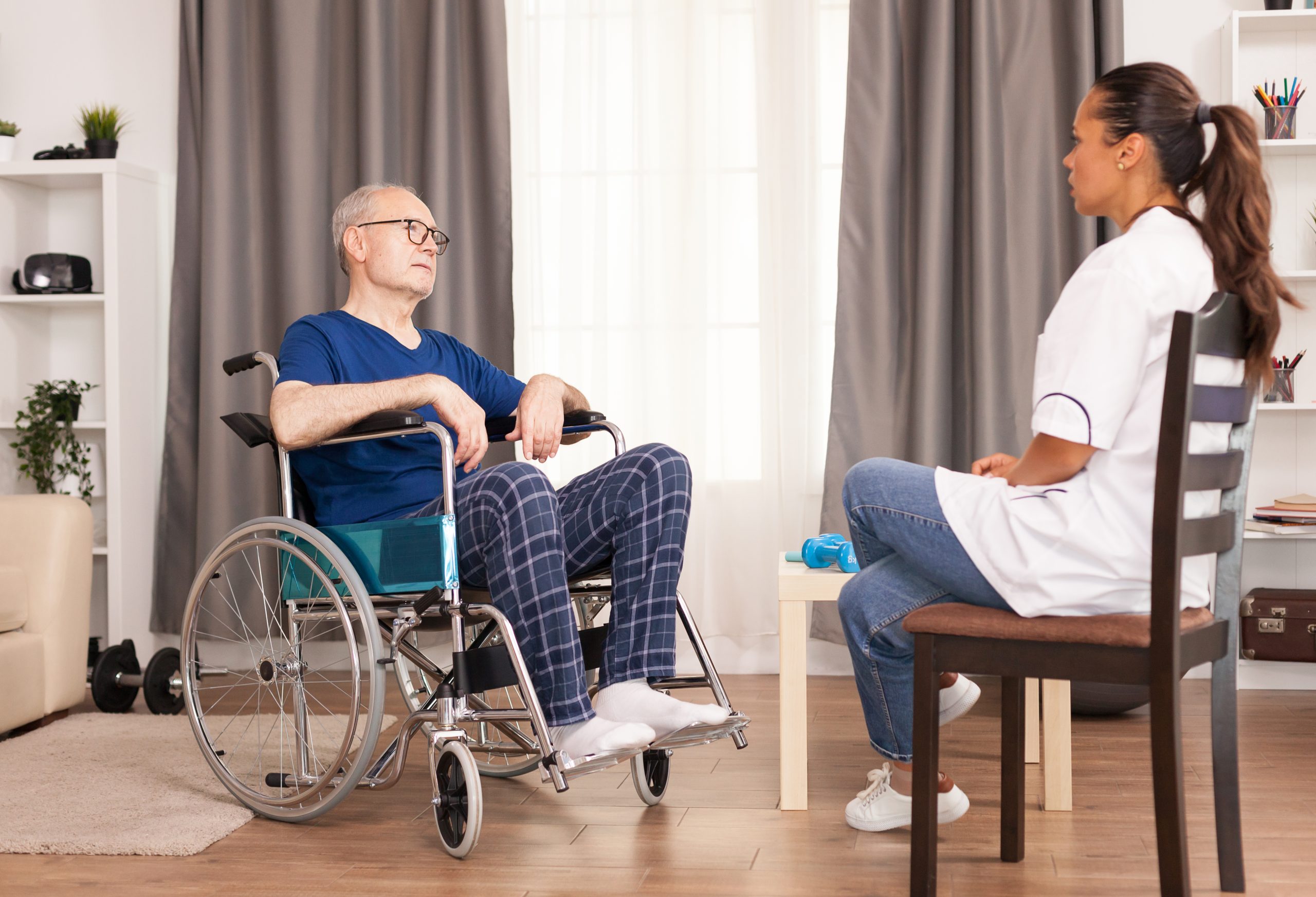 Elderly man in a wheelchair talks with a female healthcare professional seated on a chair in a living room setting.