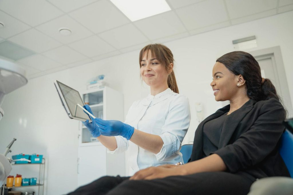 A dentist wearing gloves shows a tablet to a patient sitting in a dental chair in a brightly lit dental office.