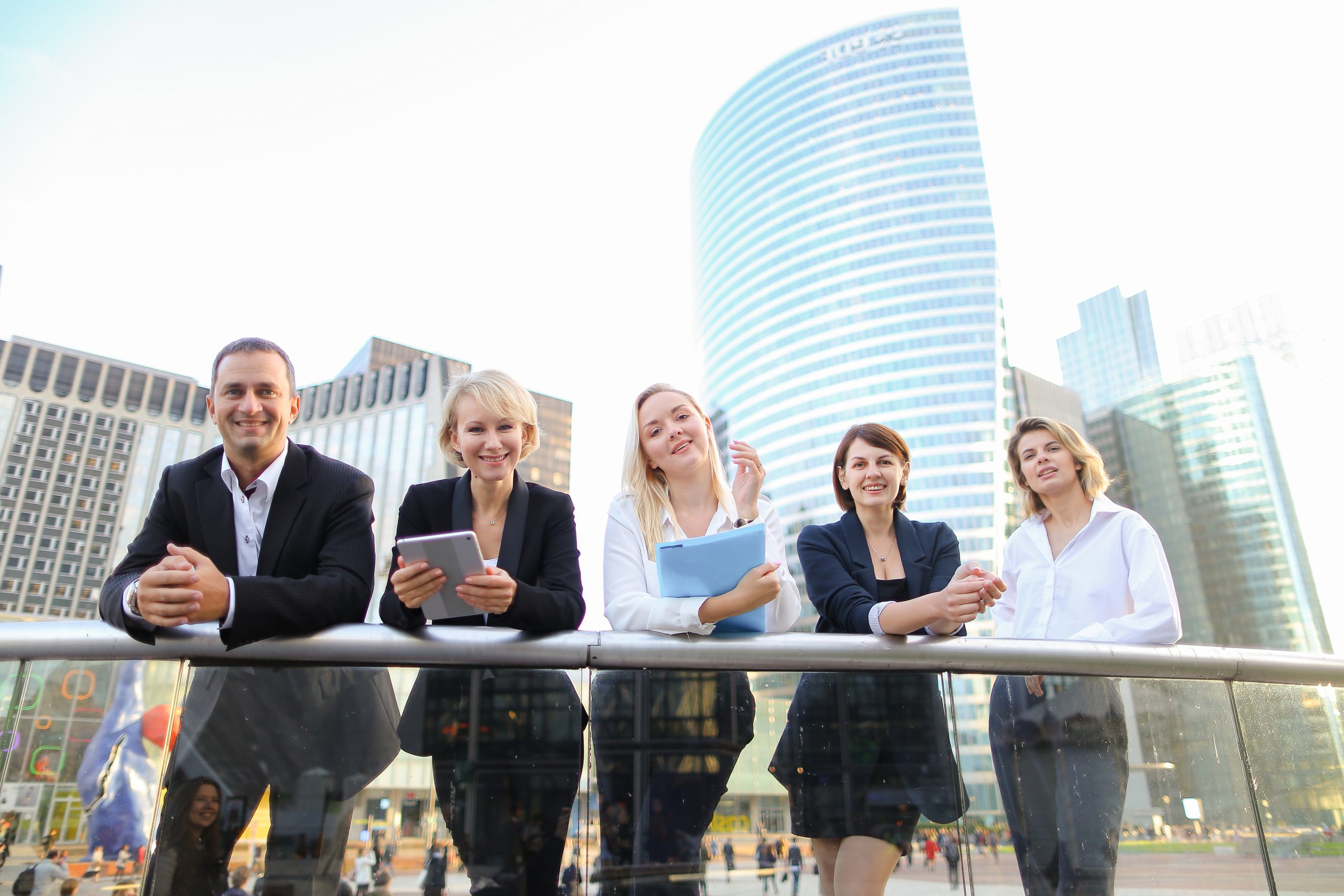 Five professionals in business attire stand by a glass railing outdoors, discussing ndis registration requirements, with modern office buildings in the background.