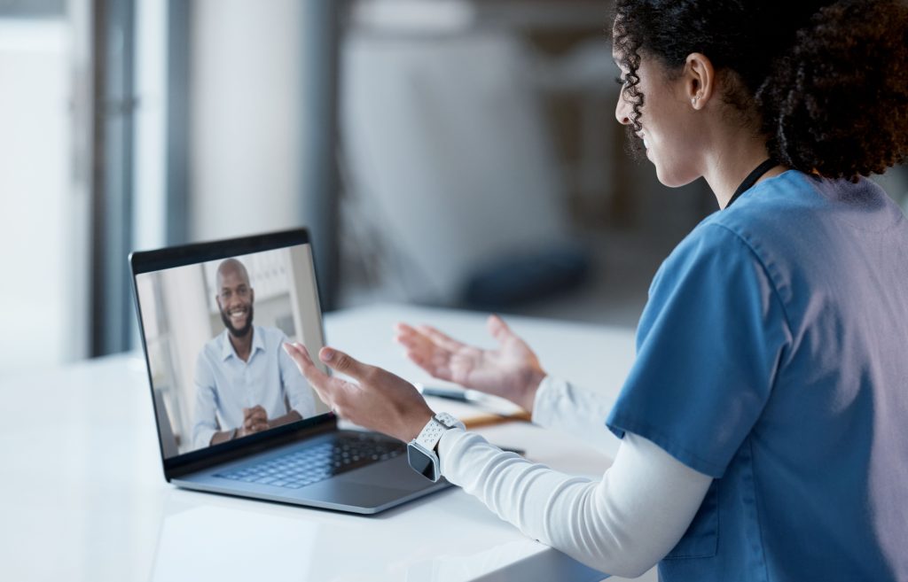 A healthcare worker in scrubs sits at a desk and talks to a man on a video call displayed on a laptop.