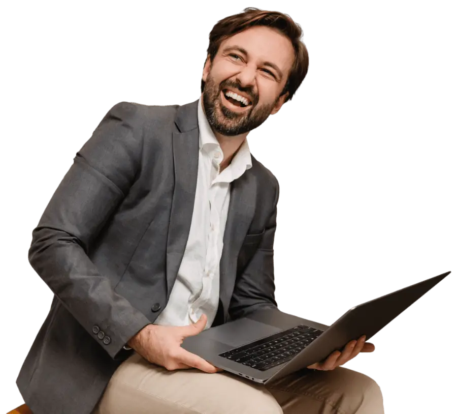 Man in a gray blazer and white shirt sitting and laughing while holding an open laptop on his lap.
