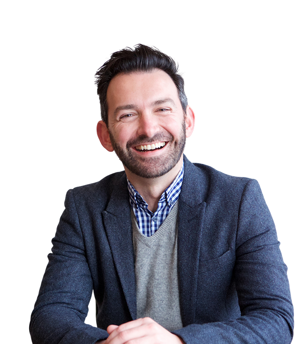 A man with dark hair and a beard, wearing a gray blazer, checkered shirt, and sweater, sits at a table and smiles at the camera against a plain background.