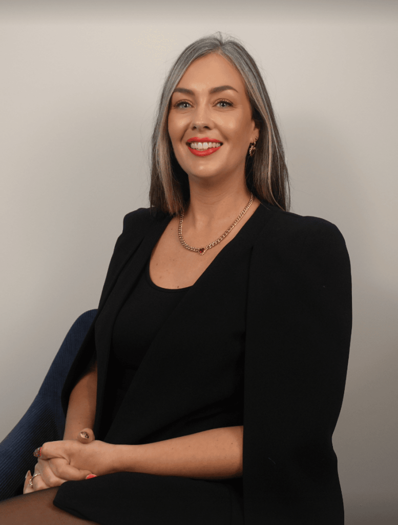 Katherine Harris, with long brown and gray hair, wearing a black outfit and gold necklace, sits on a chair against a plain background, smiling at the camera.