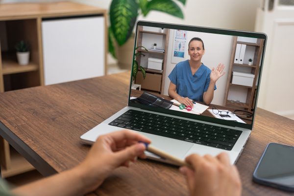 Person in scrubs waves during a video call on a laptop, while another person holding a pen sits at a desk in front of the screen.