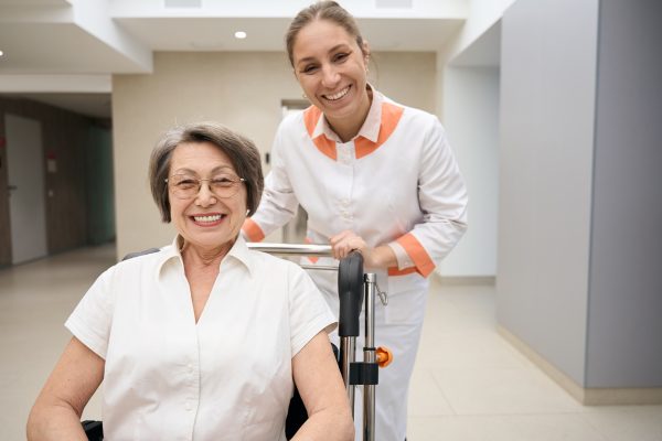 An older woman in a wheelchair smiles at the camera while a caregiver stands behind her, also smiling, in a brightly lit indoor setting.
