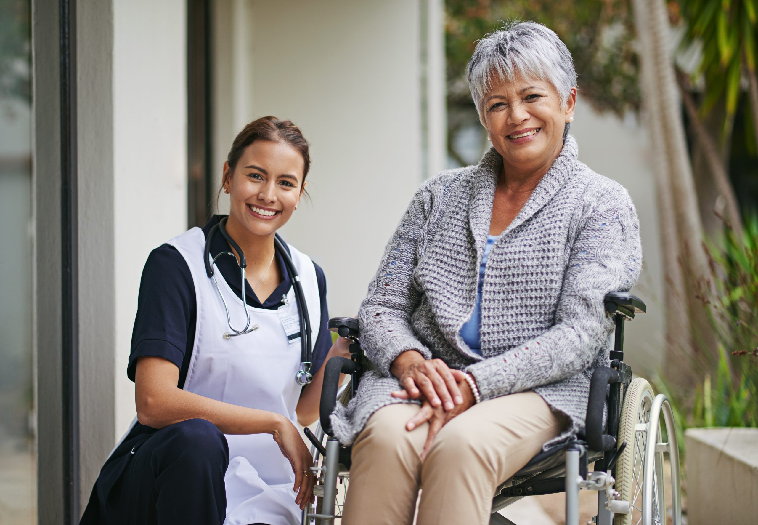 A nurse kneels beside an older woman sitting in a wheelchair outdoors; both are smiling at the camera.