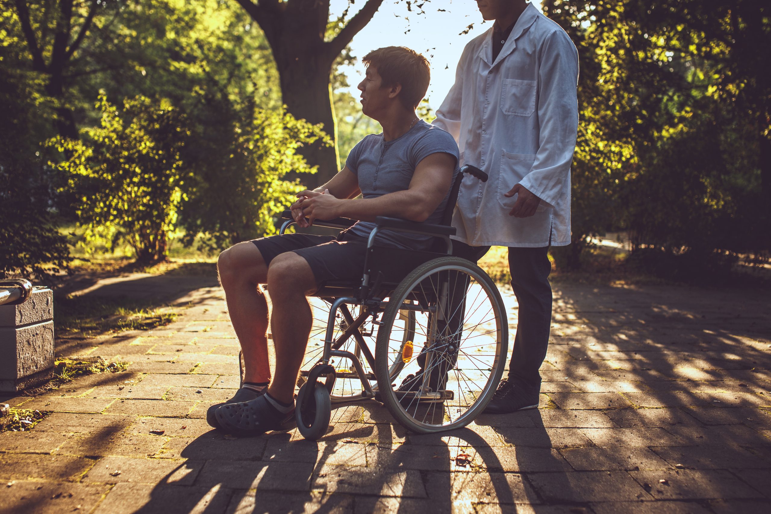 A man in a wheelchair is outdoors on a pathway, accompanied by a person in a white coat. As sunlight filters through the trees, they discuss topics like NDIS certification vs verification.