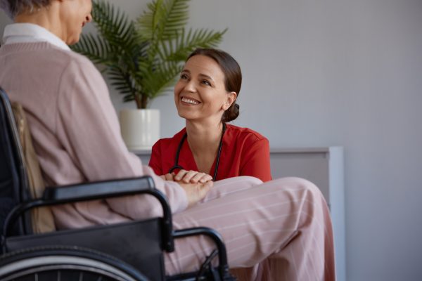 A woman in a red shirt kneels and smiles while holding hands with a person sitting in a wheelchair. A potted plant is in the background.