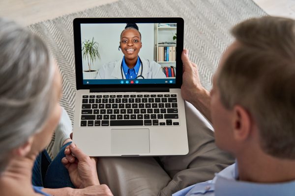 Two people sit together on a couch holding a laptop, participating in a video call with a smiling healthcare professional on the screen.