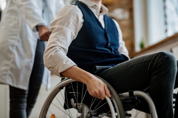 A person sits in a wheelchair, holding the wheel, while a healthcare professional stands nearby.
